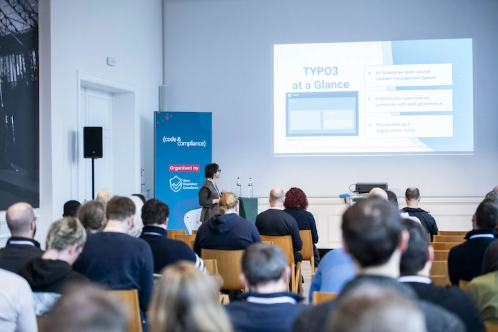 Code & Compliance Community Day 2026: A view of a room full of people listening to a presentation. The slide is titled 'TYPO3 at a Glance'.